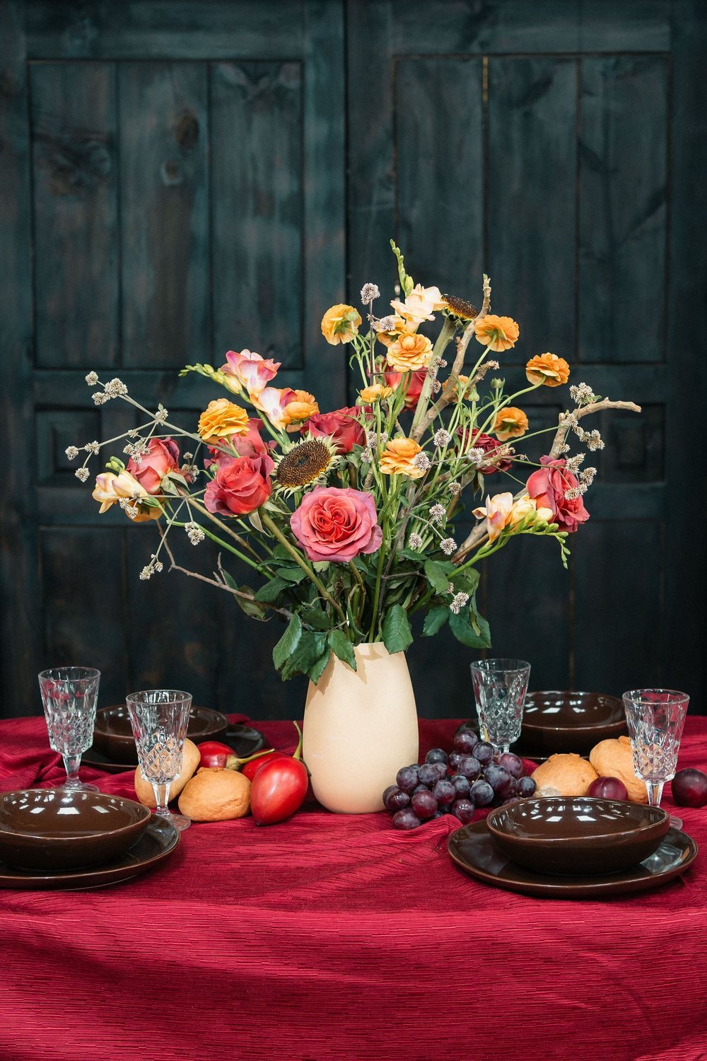 Terracotta garden roses, orange ranunculus and peach freesia in a cream vase on a table set with cocoa ceramics and deep red linen