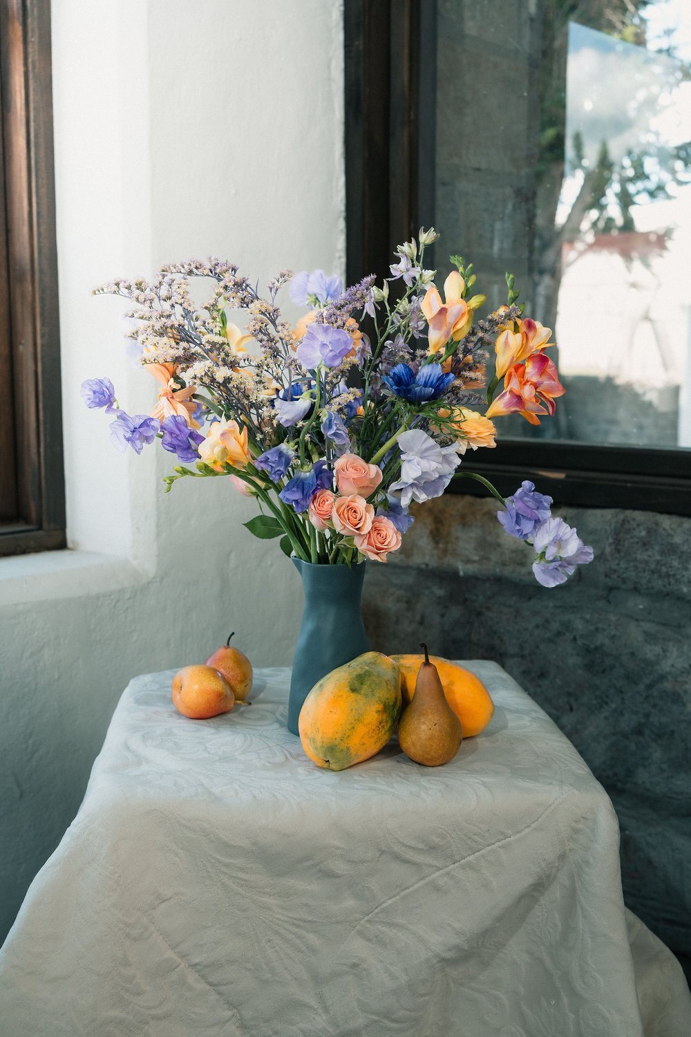 Lavender sweet peas, lilac larkspur and a blue anemone with apricot freesia in a slate vase on a white table